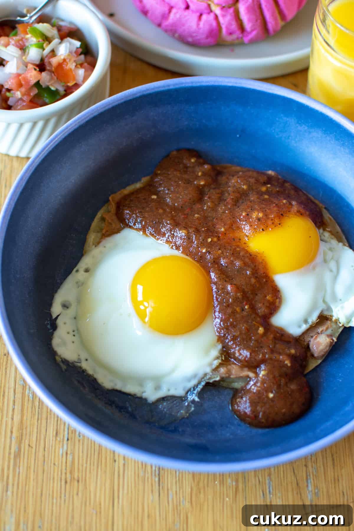 A blue plate with Huevos Rancheros in it on a wood table with a glass of orange juice next to it and a dish of Pico de Gallo. 