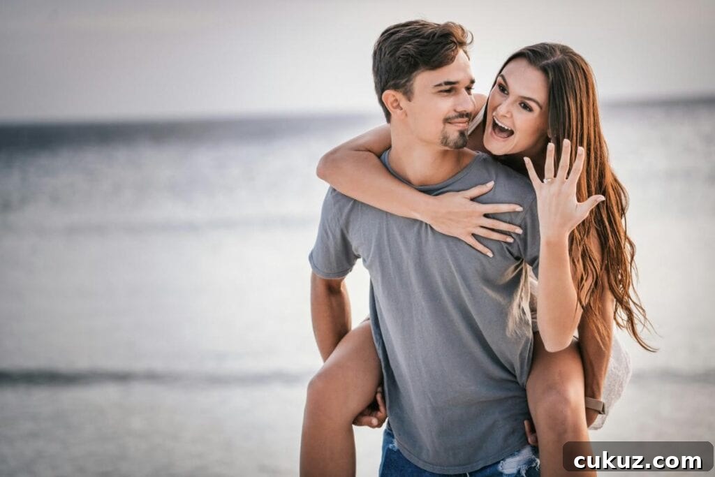 man giving woman a piggyback ride while she shows off her engagement ring on the beach