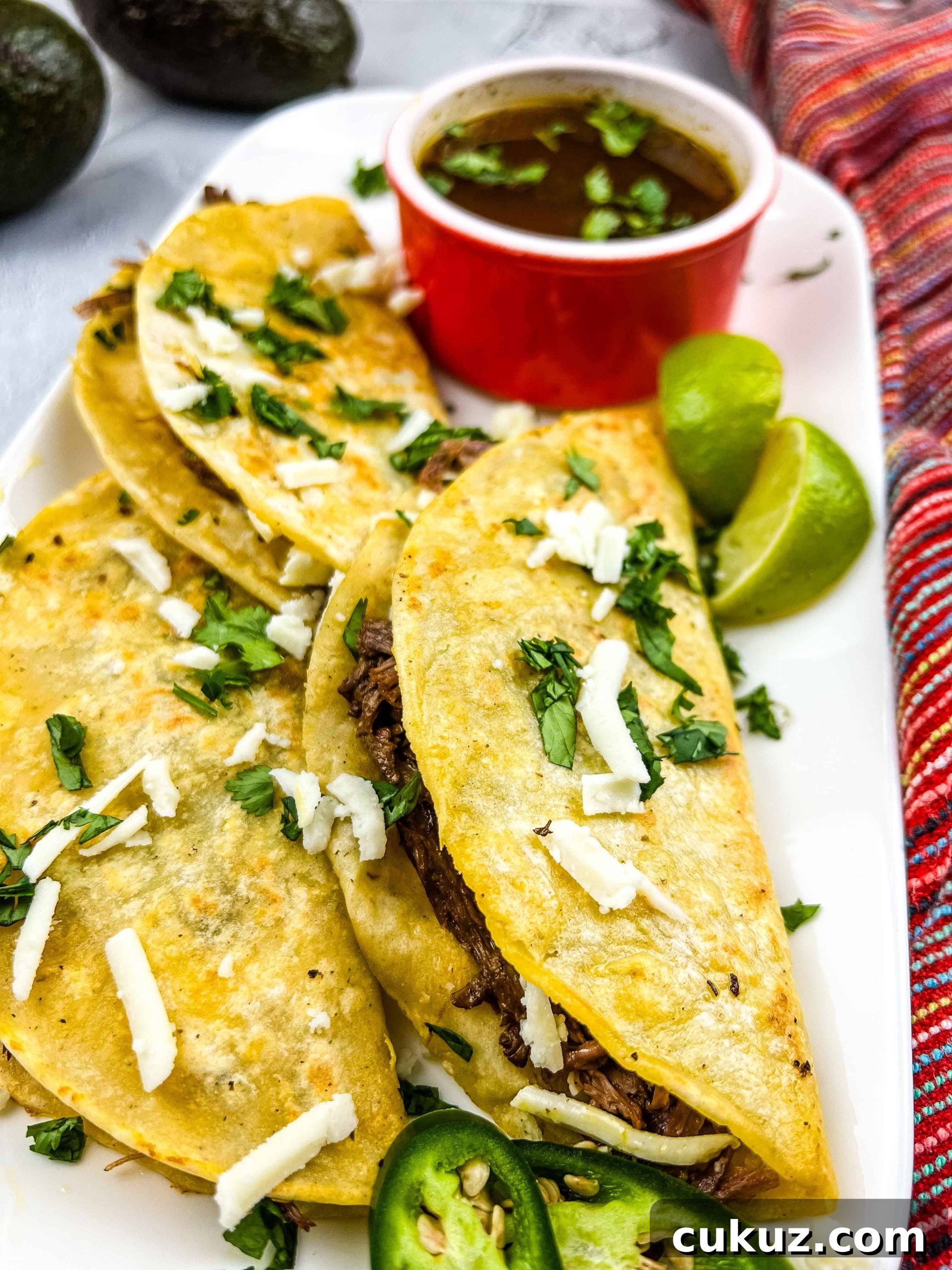 Birria Tacos Recipe with Dipping Consomme 2 birria tacos on a plate with a cup of jus in the background
