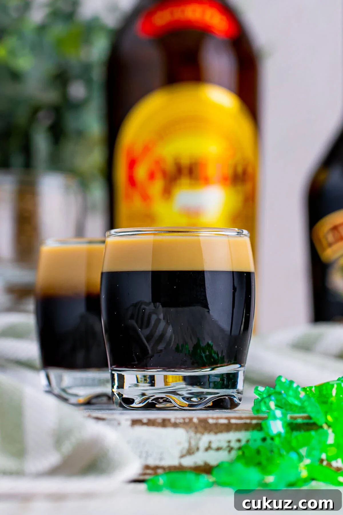 How to Make a Baby Guinness Shot: Creamy Espresso Liqueur Cocktail 2 Close-up straight on image of Baby Guinness with festive green clover necklace. Bottle of Kahlua in the background on a chipped wood serving board.