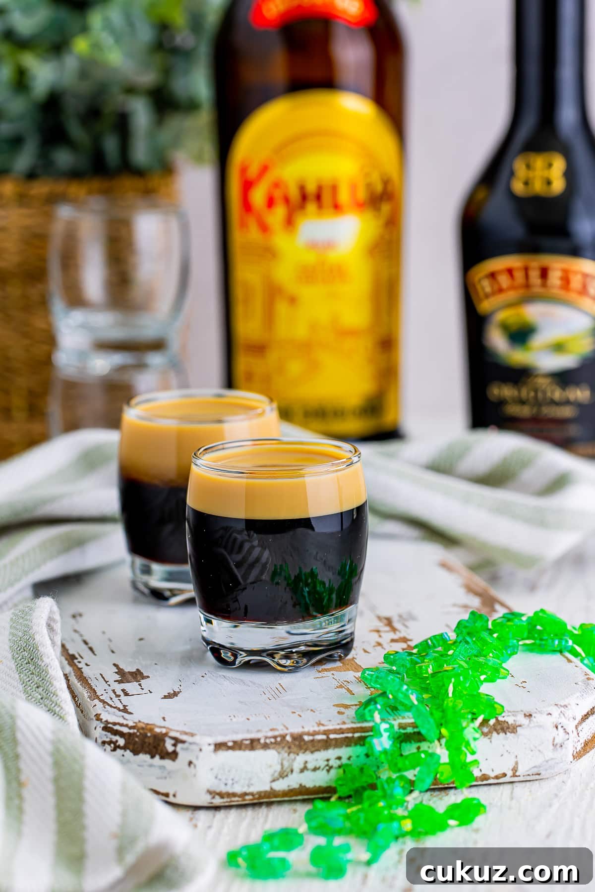 How to Make a Baby Guinness Shot: Creamy Espresso Liqueur Cocktail 3 Baby Guinness are on a white chipped wooden serving board with green festive necklace. Alcohol bottles in the background with striped green linen.