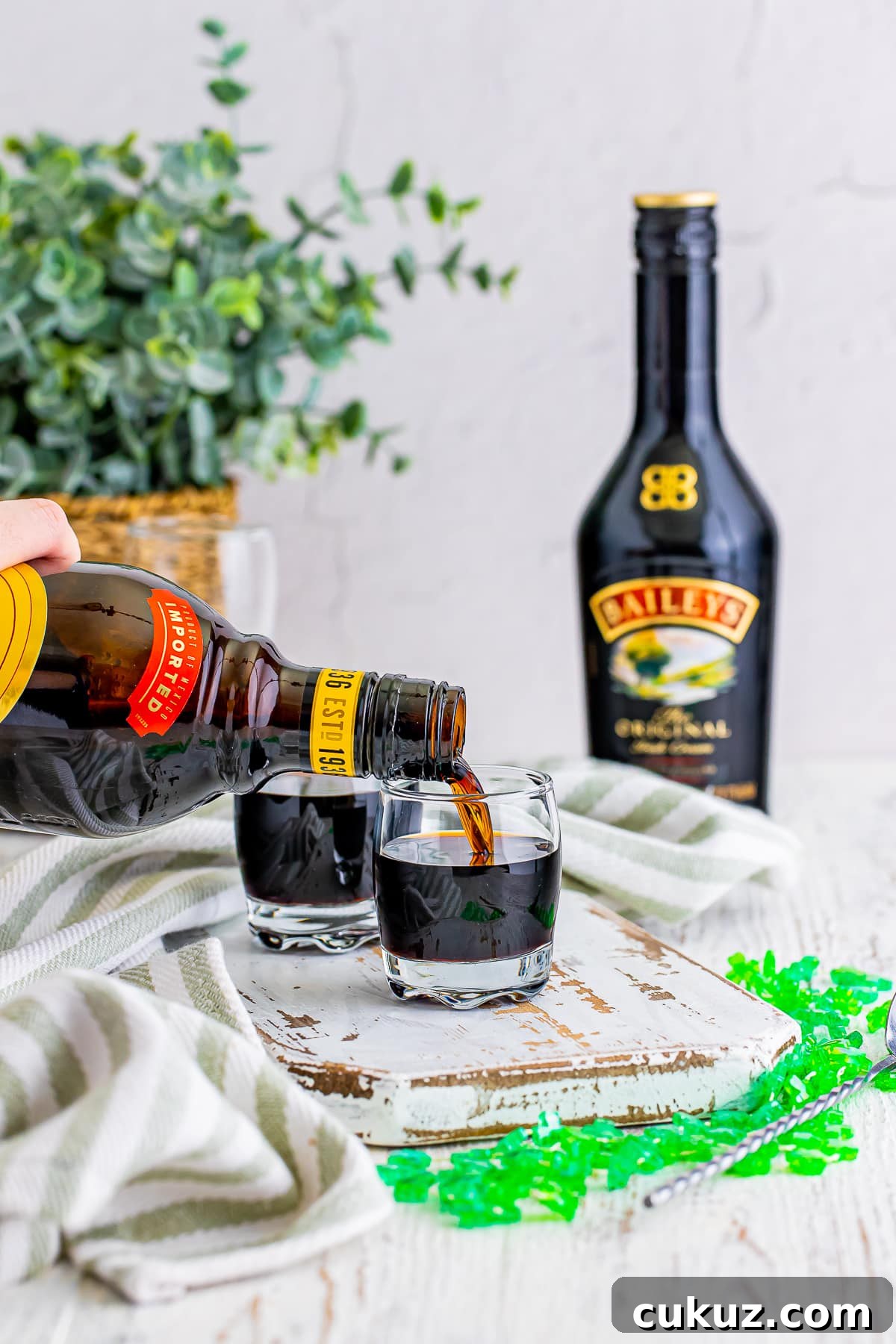 How to Make a Baby Guinness Shot: Creamy Espresso Liqueur Cocktail 5 Kahlua being poured into a shot glass for Baby Guinness Shots on a white chipped wooden serving board, green festive necklace. Plant in wicker holder in background
