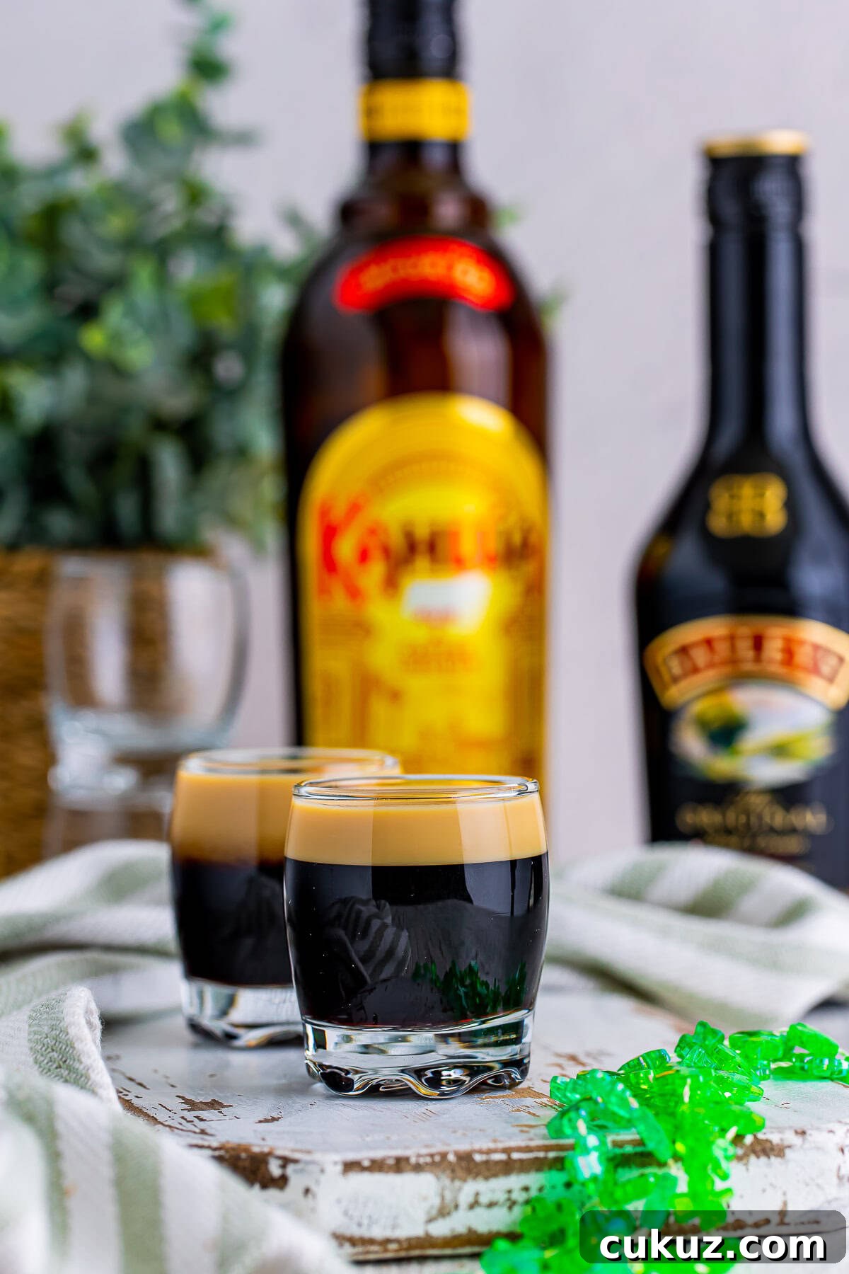 How to Make a Baby Guinness Shot: Creamy Espresso Liqueur Cocktail 7 Straight on image of Baby Guinness on a white wooden chipped serving board, festive green necklace in foreground, alcohol bottles in background. Plant in a thicker holder in background, green striped linen.