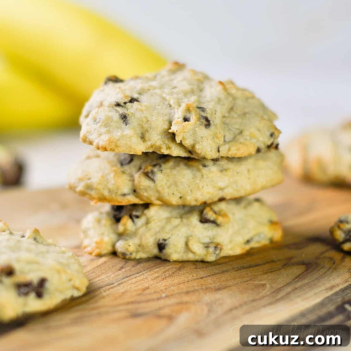 three banana bread chocolate chip cookies stacked on a cutting board.
