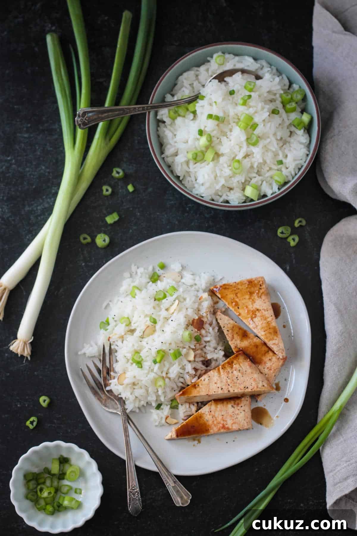 Serving of coconut jasmine rice and baked tofu on a plate next to a bowl of rice and loose green onions.