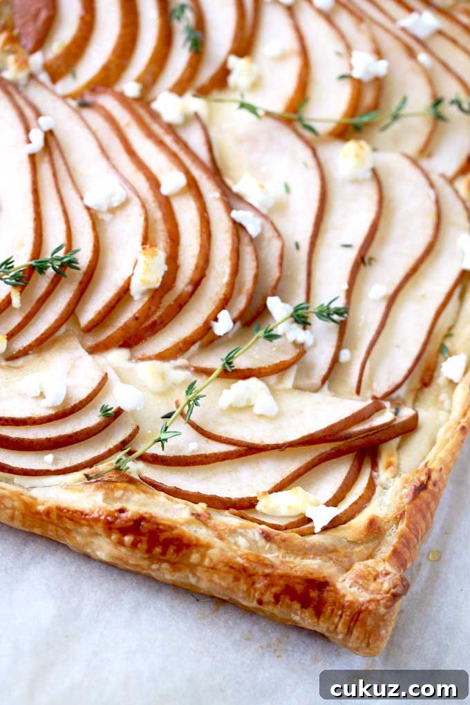 Pear and Goat Cheese Tart with Honey and Thyme 2 View of a golden brown puff pastry tart topped with thinly sliced pears garnished with goat cheese and fresh thyme on a parchment paper lined baking sheet.