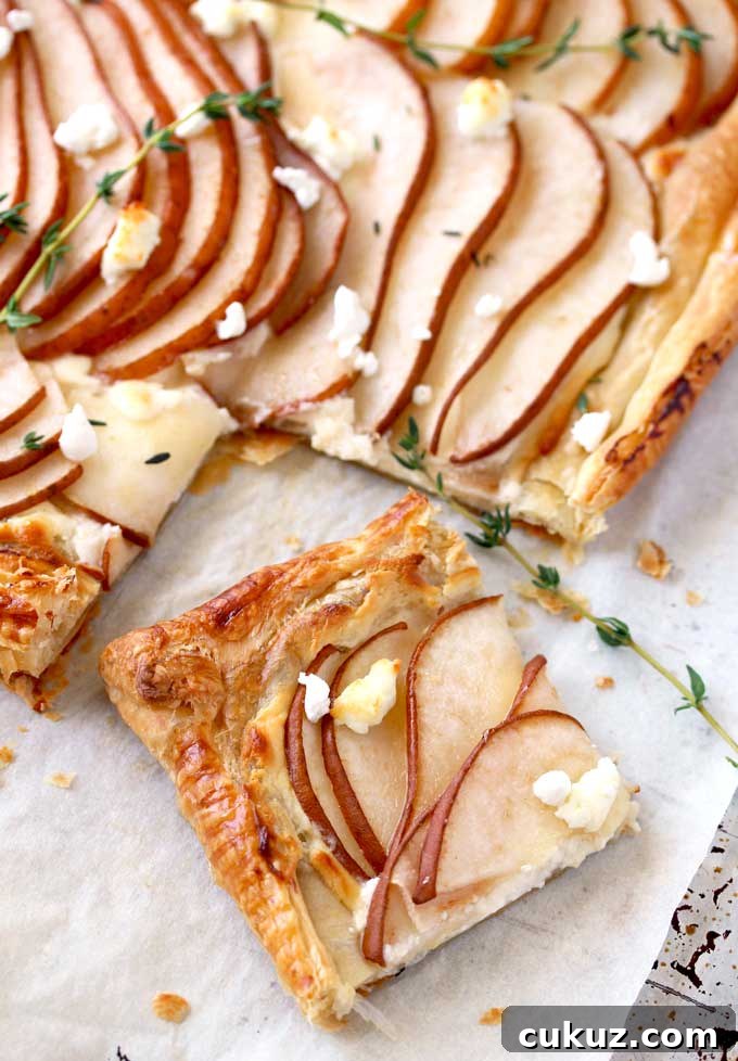 Pear and Goat Cheese Tart with Honey and Thyme 3 View of a golden brown puff pastry tart sliced The tart is topped with thinly sliced pears garnished with goat cheese and fresh thyme on a parchment paper lined baking sheet.