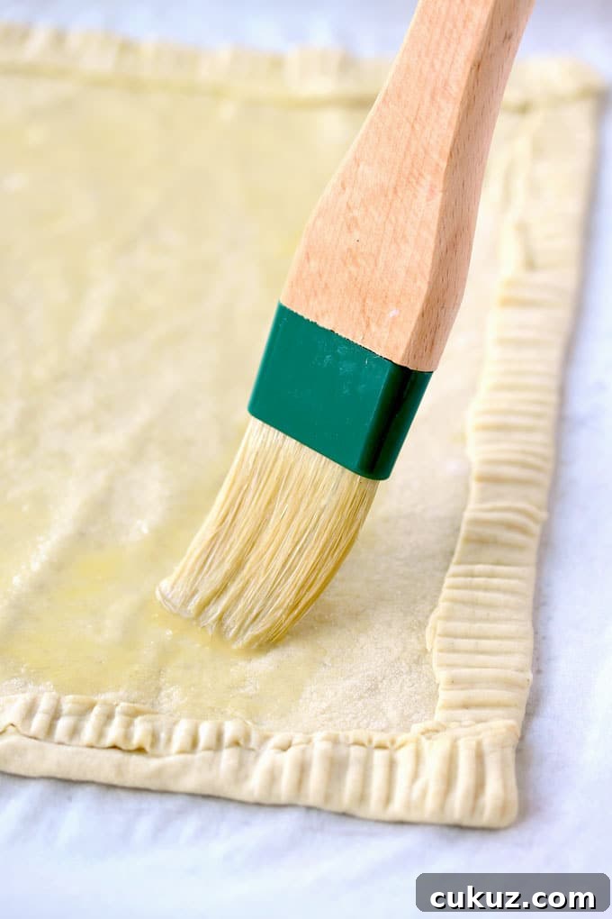 Pear and Goat Cheese Tart with Honey and Thyme 4 View of a sheet puff pastry rolled out. The edges have been fold to create a border and crimped. You can see a pastry brush brushing the dough with an egg wash mixture.