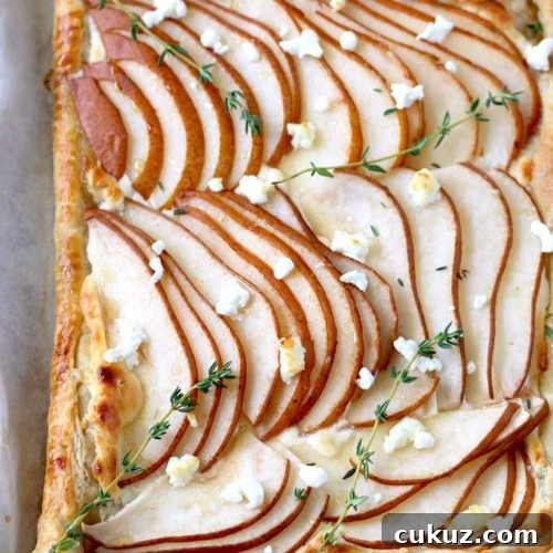 Pear and Goat Cheese Tart with Honey and Thyme 7 Top view of a golden brown puff pastry tart topped with thinly sliced pears garnished with goat cheese and fresh thyme on a parchment paper lined baking sheet.