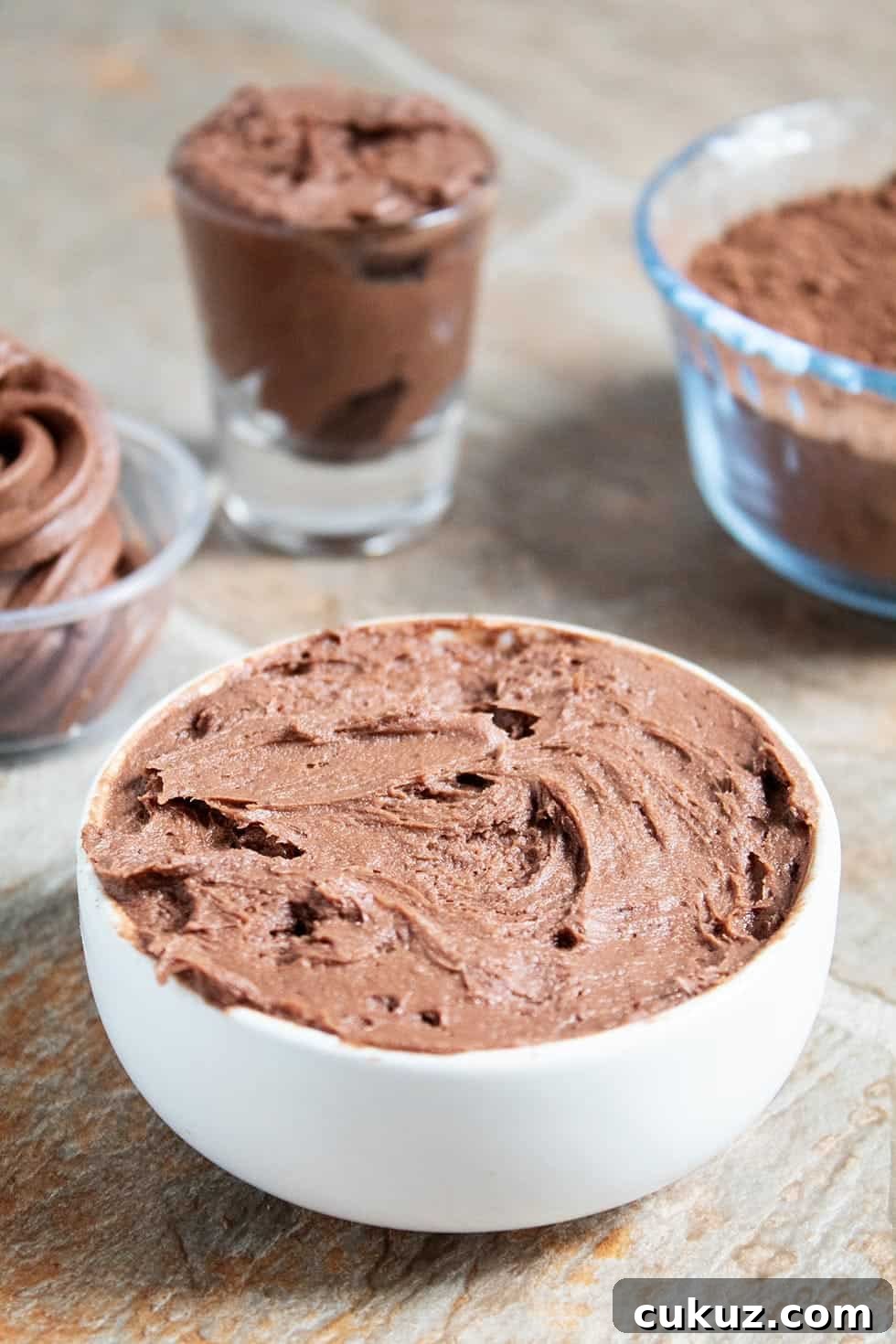 side view of white bowl containing keto chocolate frosting in front of other bowls and cocoa powder
