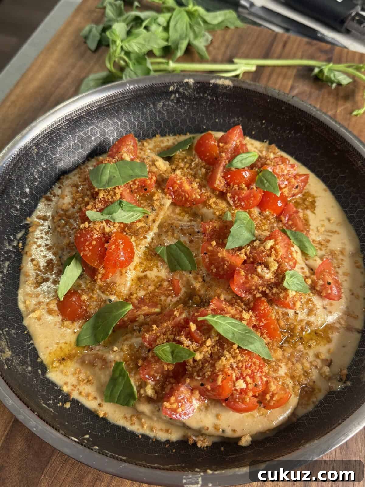 Crispy Baked Chicken Parm with Light Marinara 3 A skillet with creamy sauce topped with chopped cherry tomatoes, fresh basil leaves, and golden breadcrumbs sits on a wooden surface, with sprigs of fresh basil nearby.