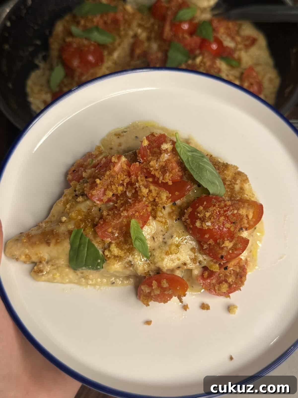 Crispy Baked Chicken Parm with Light Marinara 4 A plate with a piece of chicken breast topped with halved cherry tomatoes, fresh basil leaves, and crispy breadcrumbs, with more of the dish visible in the background.
