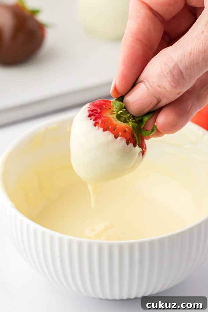 A strawberry that has been dipped in melted white chocolate being held over the bowl of chocolate, letting the excess chocolate drizzle off