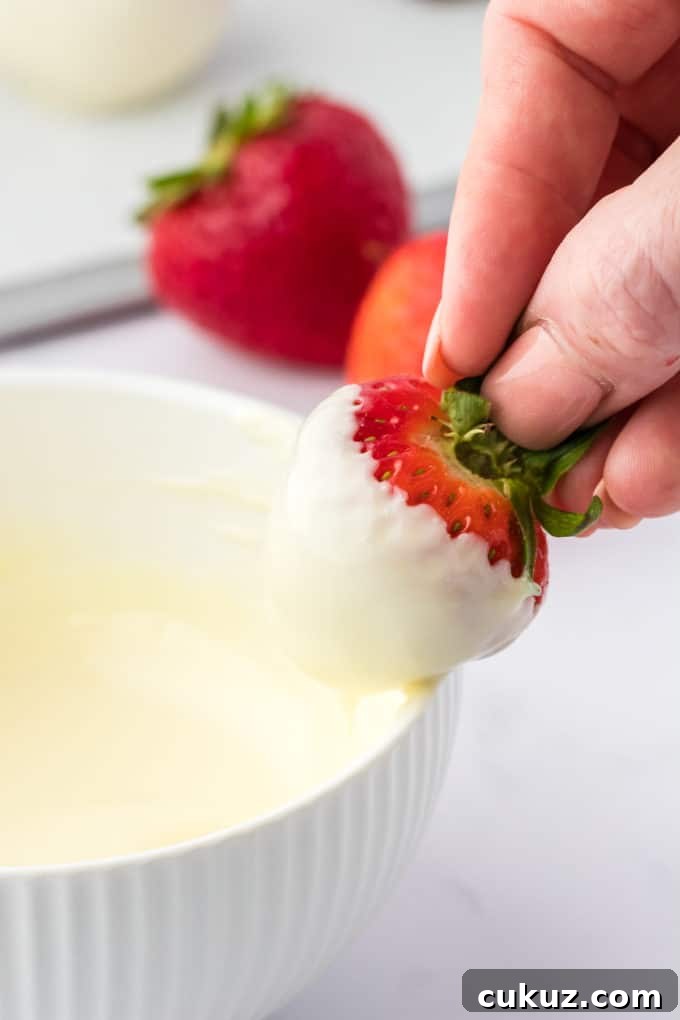 A strawberry that has been dipped in melted white chocolate being held over the bowl of chocolate, letting the excess chocolate drizzle off