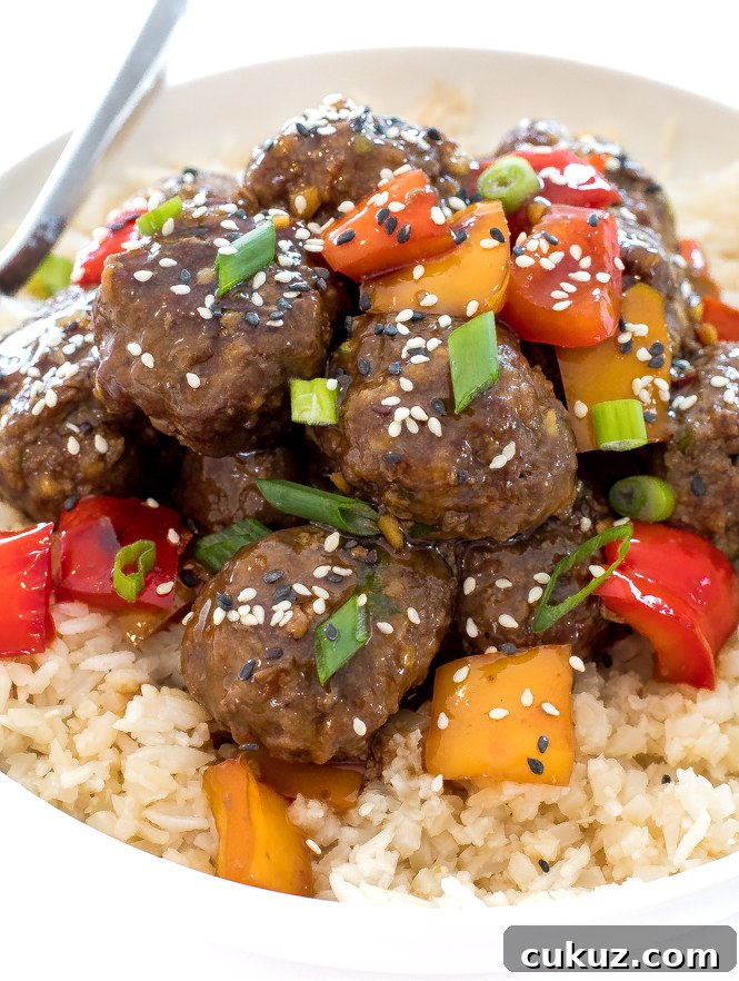 Teriyaki style meatballs in a large white bowl of white rice. Topped with sesame seeds, green onions and colorful bell pepper chunks.