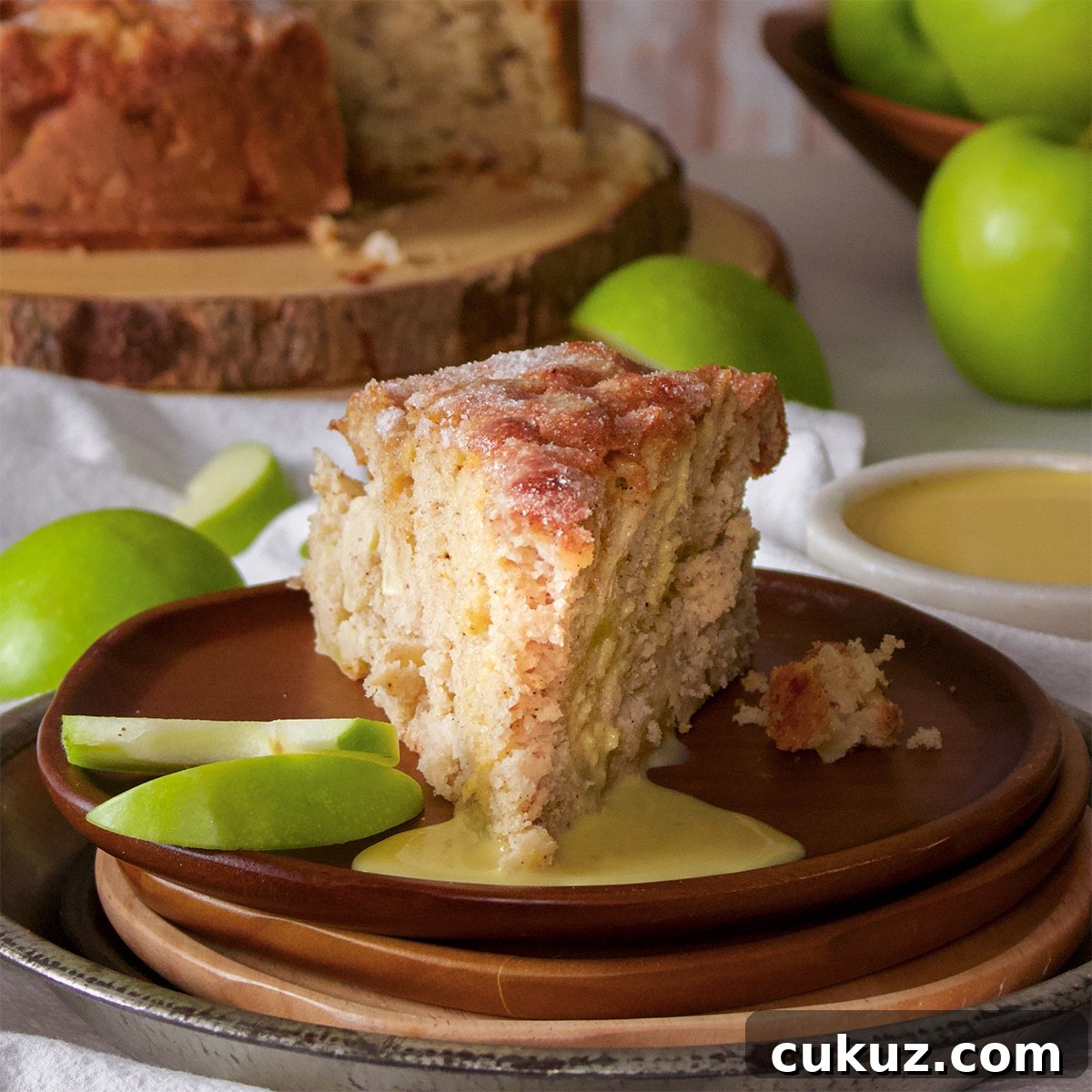 A slice of Irish Apple Cake covered in custard sauce on a wood plate with a few sliced green apples.