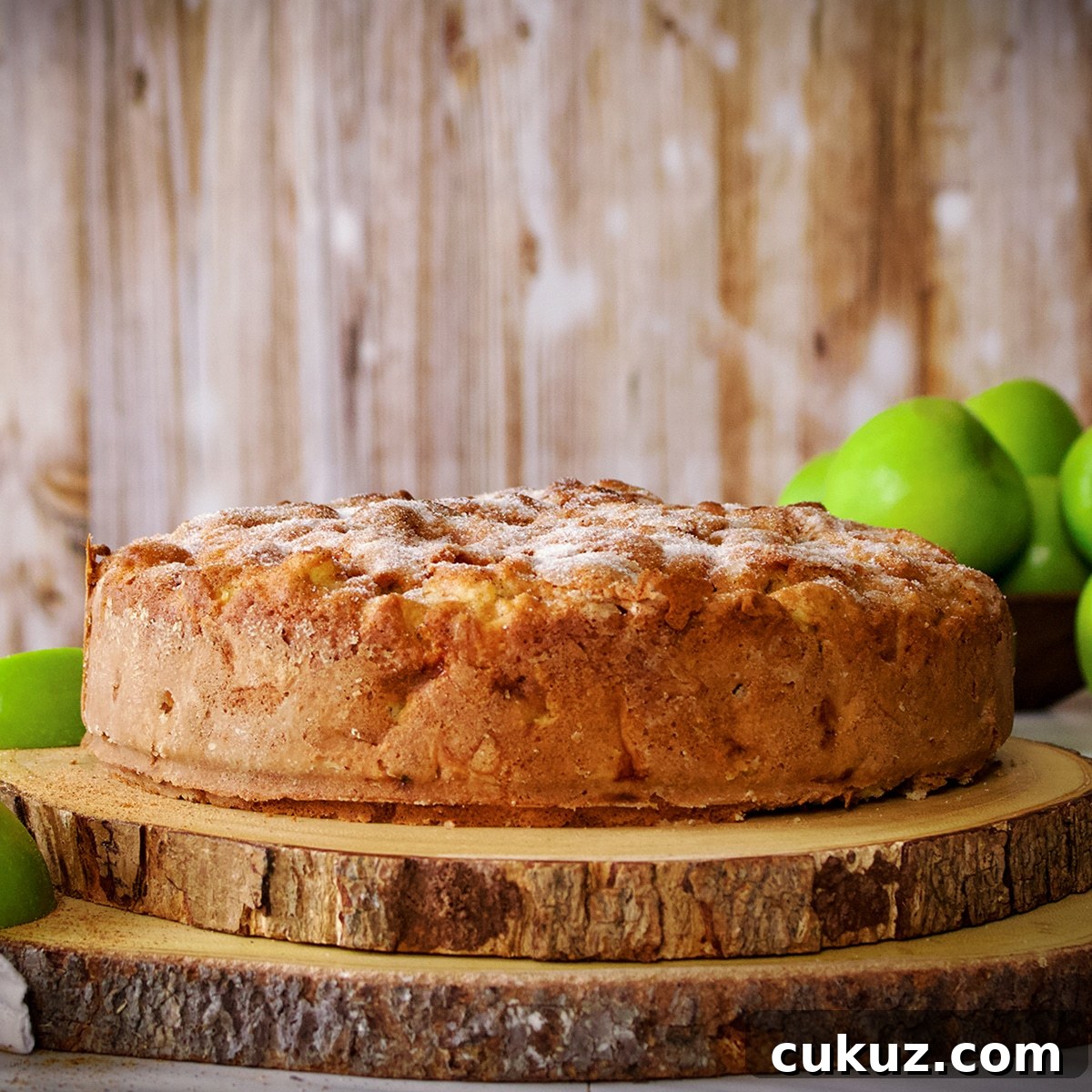 An Irish Apple Cake with a sugar crusted top on two wood cake boards surrounded by green apples.