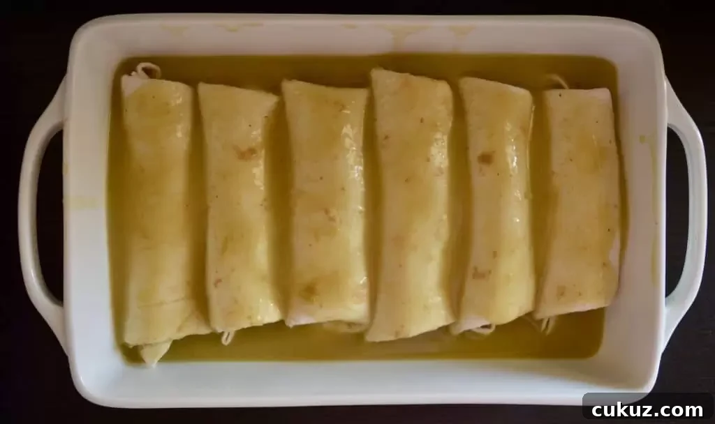 green chili enchilada sauce poured over top of six burritos in a white baking dish.