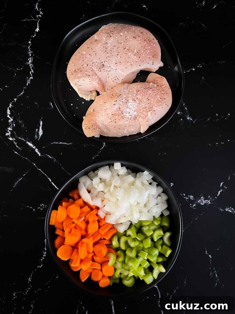 Chicken breasts on a dark plate seasoned with salt and pepper and a dark bowl full of diced onions, carrots, and celery.