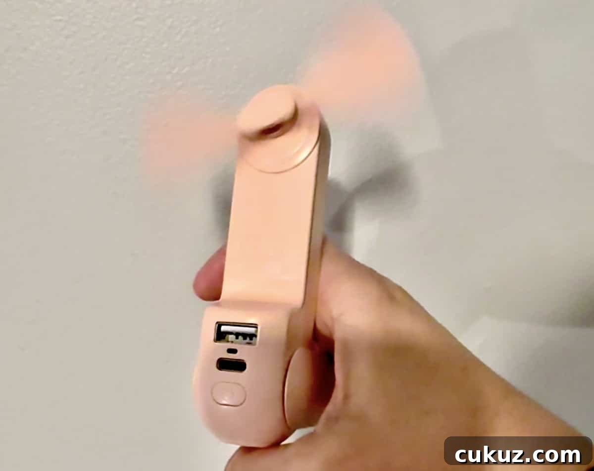 A hand holding a small, pink handheld fan with a USB port and a micro-USB charging port. The fan blades are spinning.