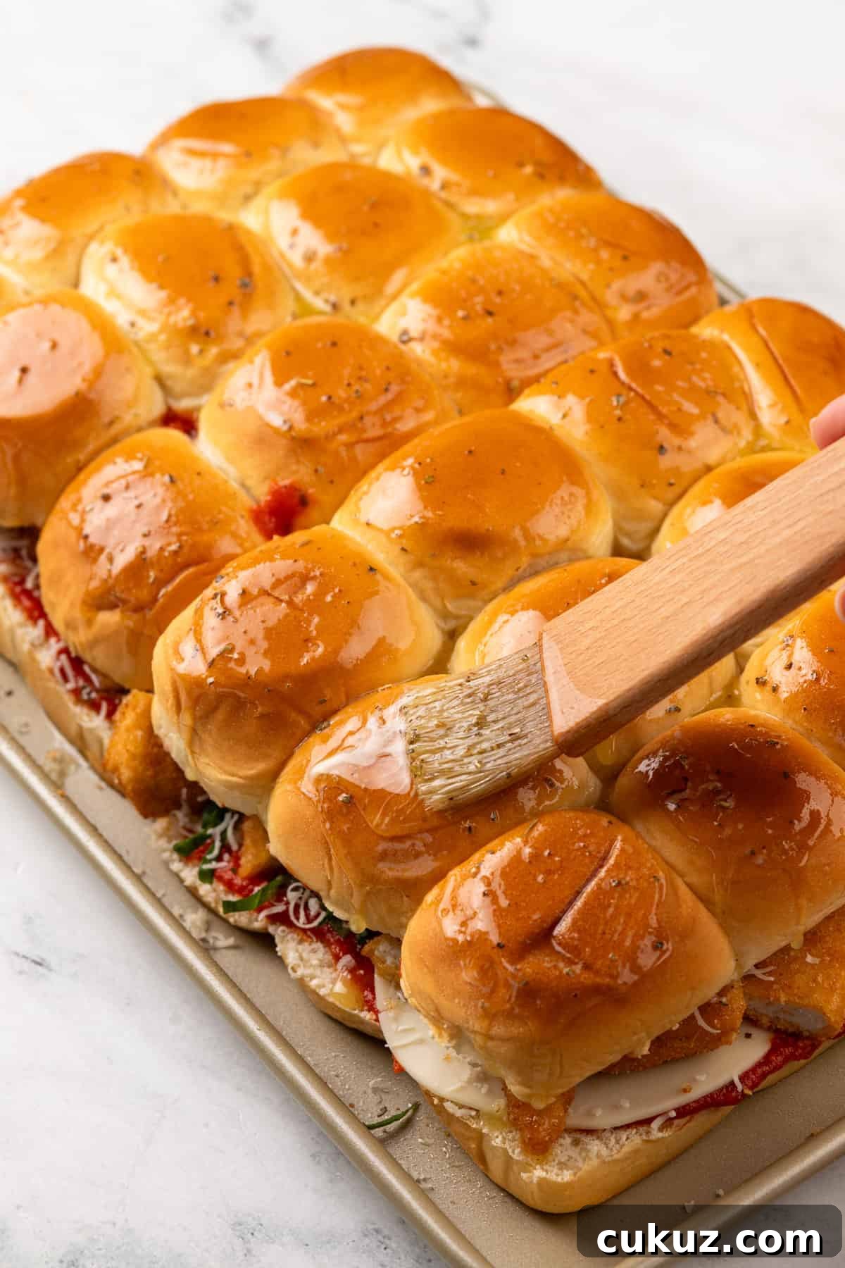 Brushing melted butter over the tops of assembled chicken parmesan sliders on a rimmed baking sheet.