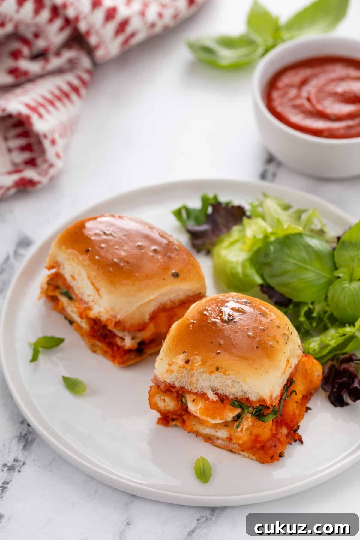 Two chicken parmesan sliders next to a green salad on a white plate.