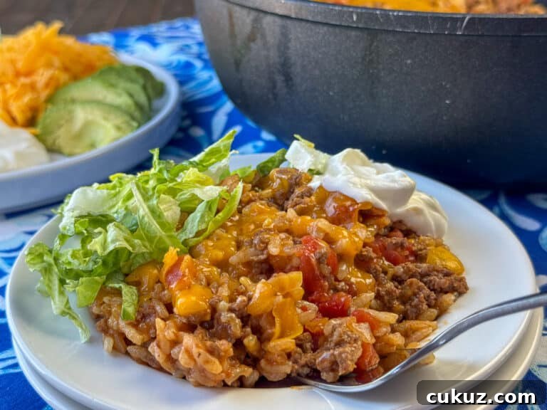 One-Pan Taco Rice Skillet Recipe for Quick Weeknight Dinners 2 Fork ready to scoop up beef and rice mixture with cheese on top and lettuce and tomato in the background with the skillet and a plate of other garnishes like avocado.