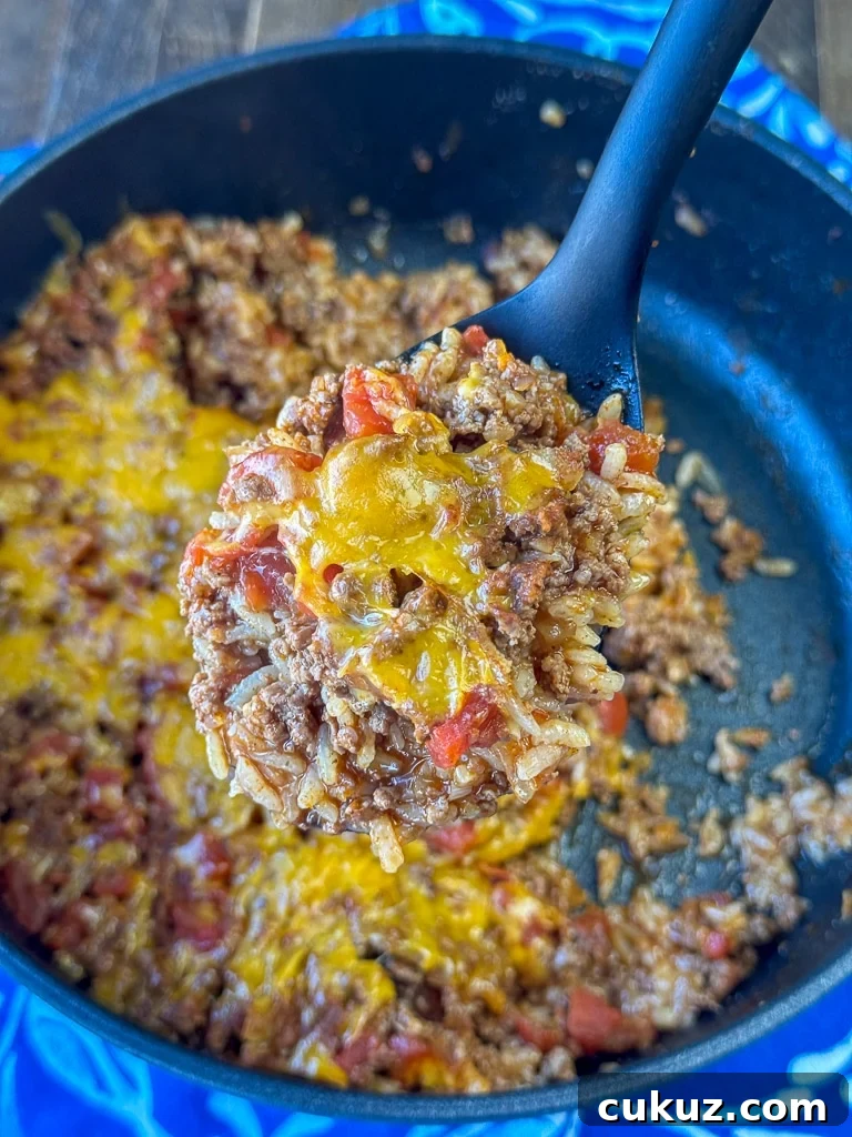 One-Pan Taco Rice Skillet Recipe for Quick Weeknight Dinners 3 Yellow cheese melted over a ground beef skillet meal with rice, tomatoes and taco seasoning on a large black spoon over top of a skillet.