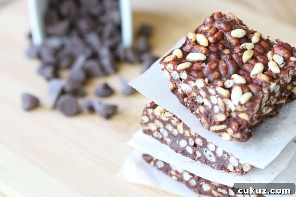 three chocolate crunch bars stacked on parchment paper with more bars, a glass of milk and a ramekin of chocolate chips behind it