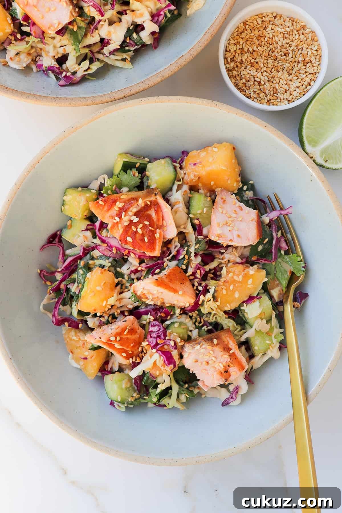 The salmon mango salad in two bowls with a fork. Lime wedge and sesame seeds are are on the side.