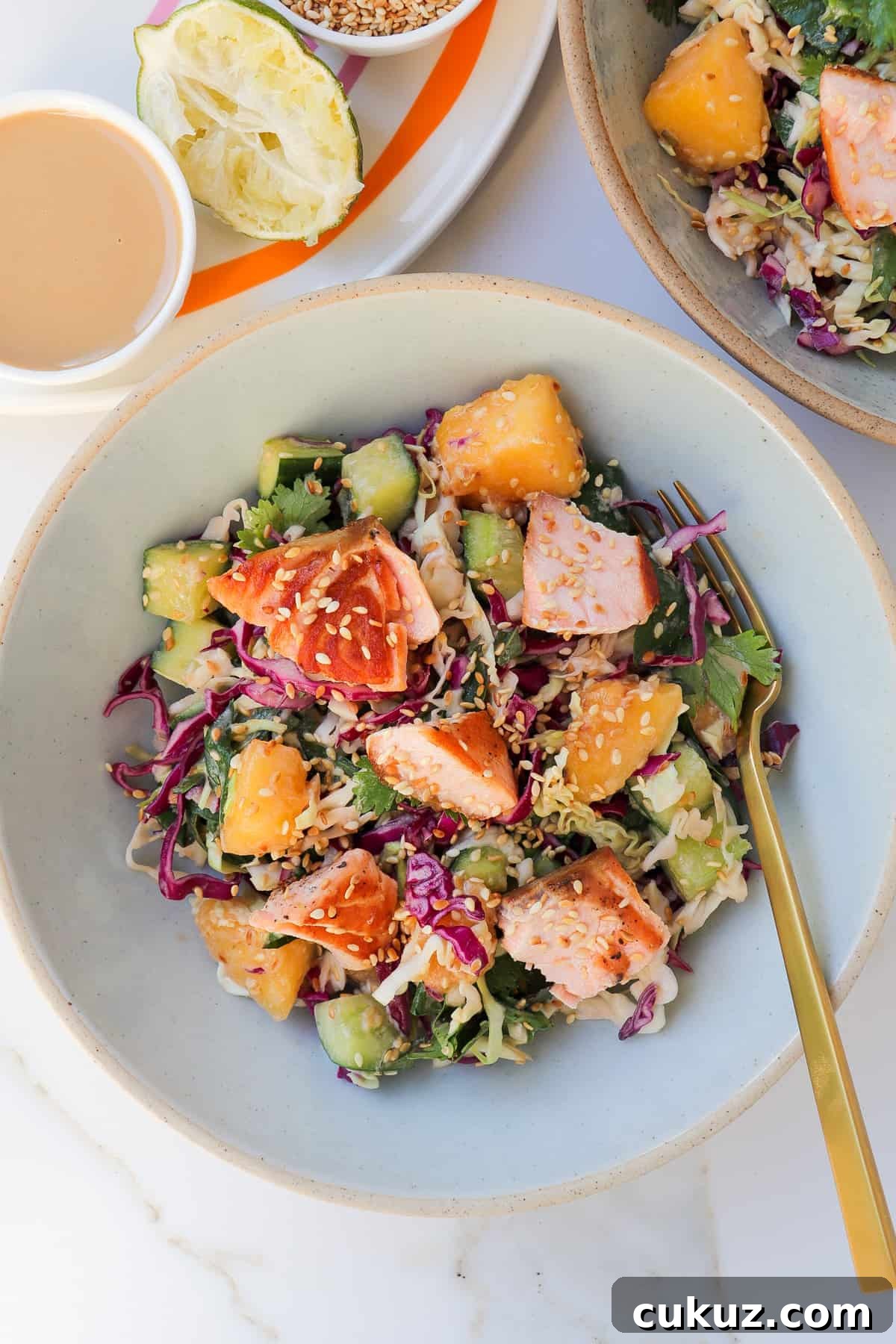 The salmon salad in two bowls with a fork. A squeezed lime wedge and tahini sauce are in a dish on top on an orange plate on the left side.