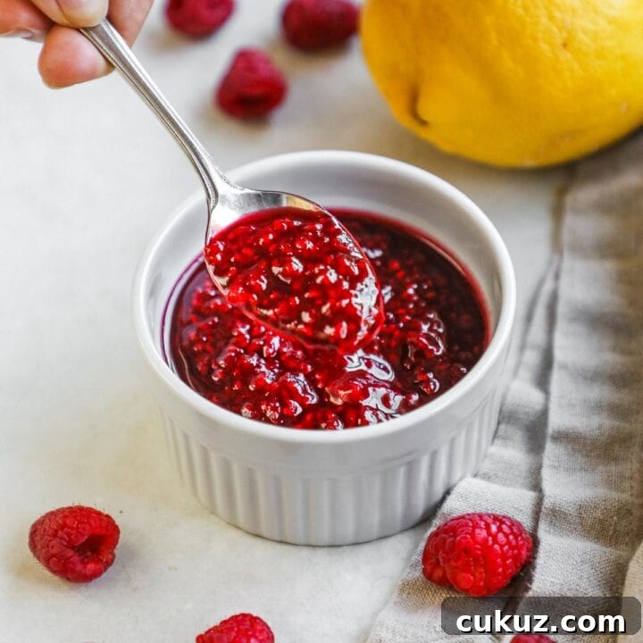 Spoon holding a scoop of handmade raspberry compote above a ramekin of compote for ice cream, cake, cheesecake, dessert, and more.