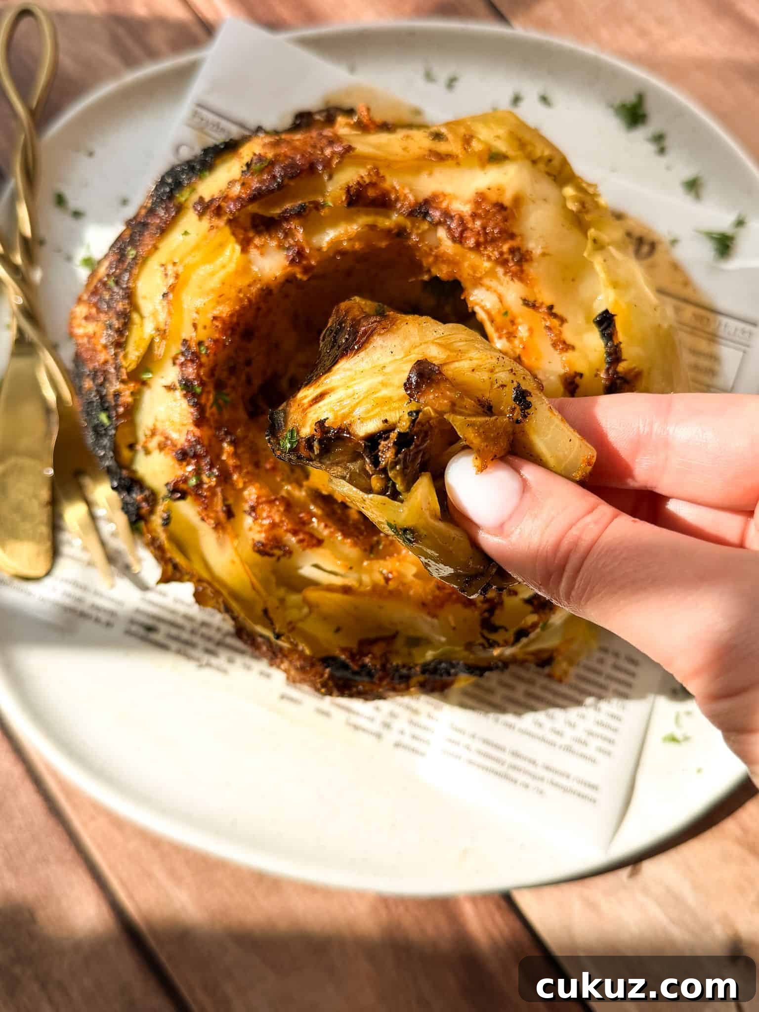 Viral Cabbage Boil Recipe: How to Make the Trendy Cabbage Pot 3 A hand holds a roasted cabbage leaf above a plate with a whole, charred roasted cabbage, garnished with herbs. A fork and knife rest on the plate, and sunlight highlights the food.
