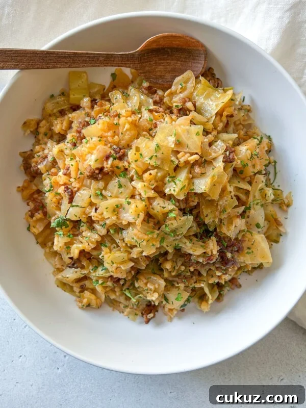 Viral Cabbage Boil Recipe: How to Make the Trendy Cabbage Pot 4 A white bowl filled with a Deconstructed Cabbage Rolls mixture of cooked cabbage and ground meat, garnished with chopped herbs, sits on a light surface with a wooden spoon resting on the rim. A beige cloth is partially visible beside the bowl.