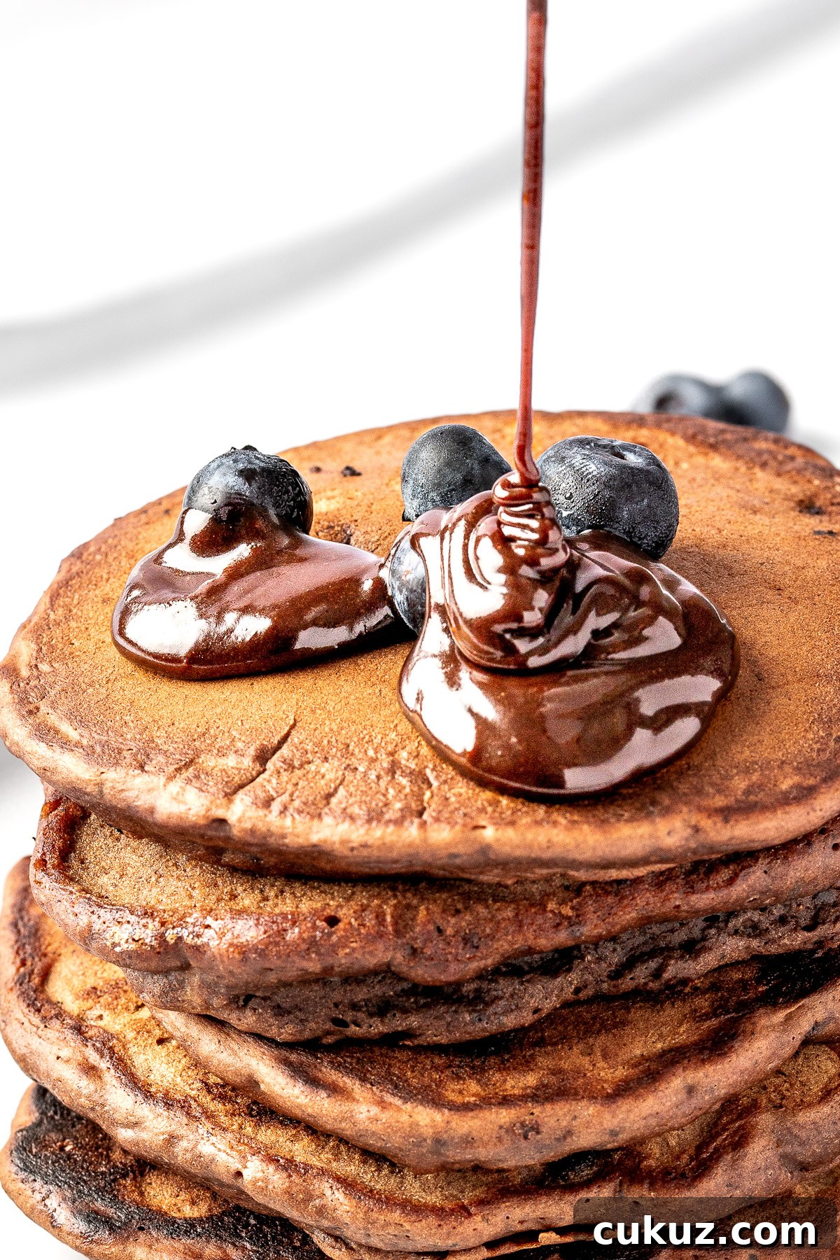 Chocolate pancake syrup being drizzled on top of a stack of pancakes.