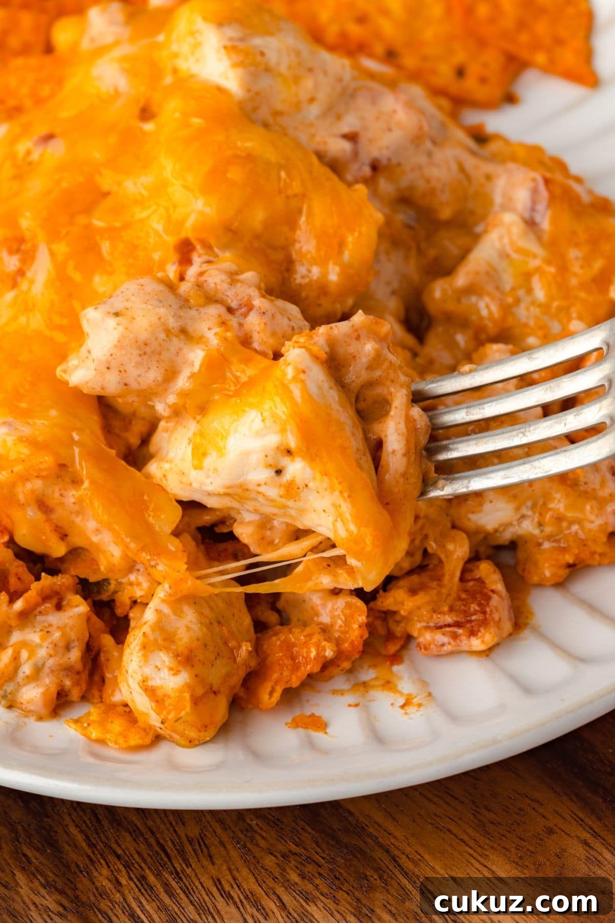 Close-up of a cheesy chicken casserole on a white plate. A fork is lifting a piece, revealing layers of melted cheese and chunks of chicken mixed with a creamy sauce. The dish has a golden-brown crust.