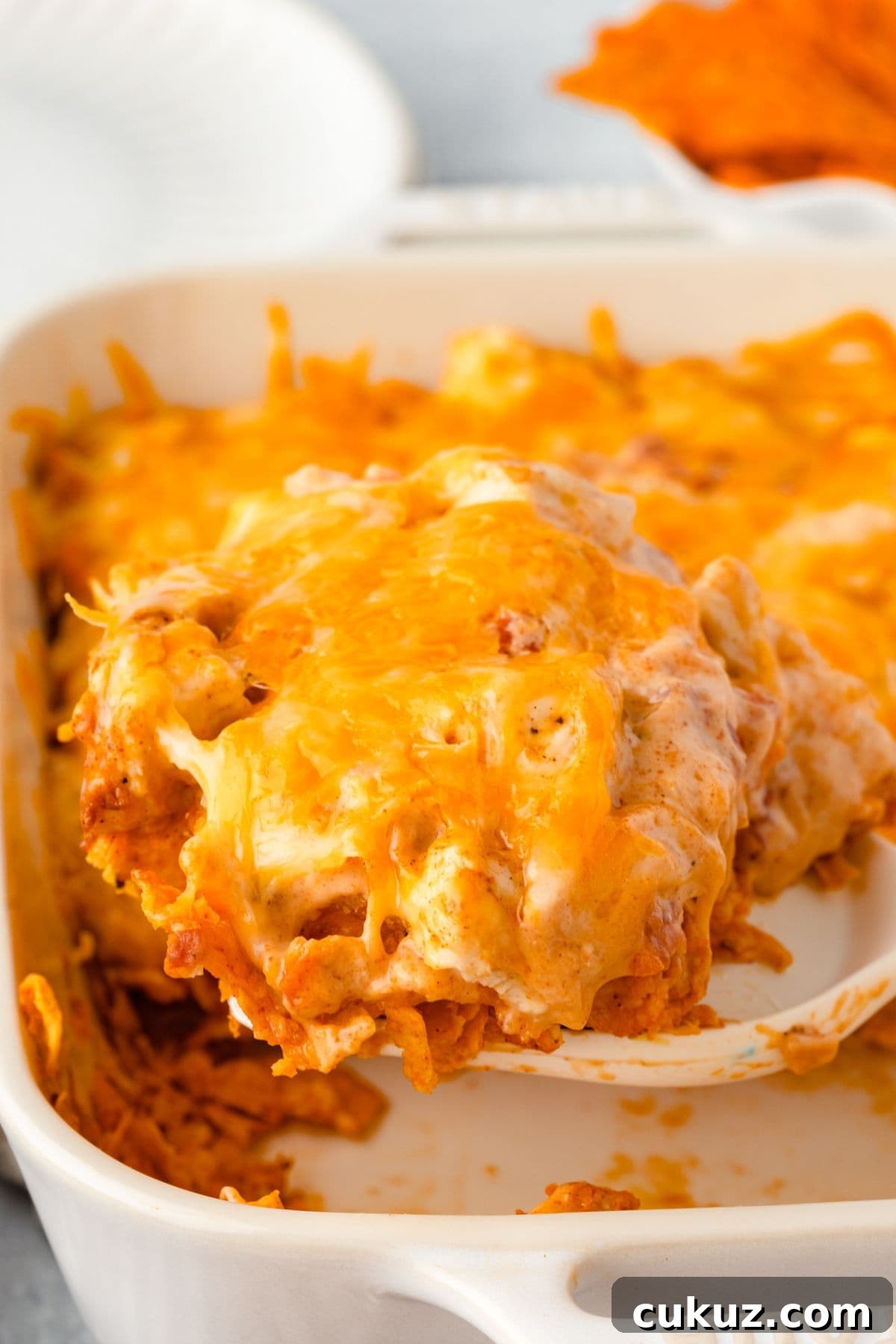 A close-up of a cheesy tortilla chip casserole in a white baking dish. A slice is being lifted, revealing layers of melted cheese and crunchy chips. The background has an orange napkin and another part of the dish visible.