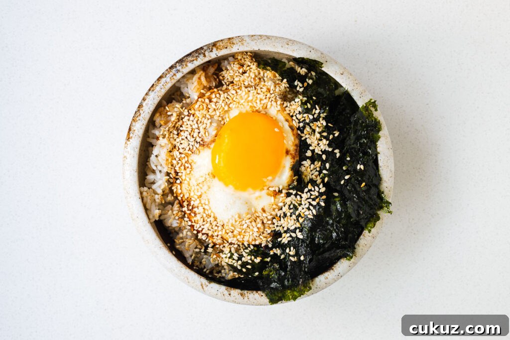 An assembled gyeran bap breakfast bowl with buttered rice, fried egg, roasted seaweed and toasted sesame seeds drizzled with soy sauce and sesame oil.