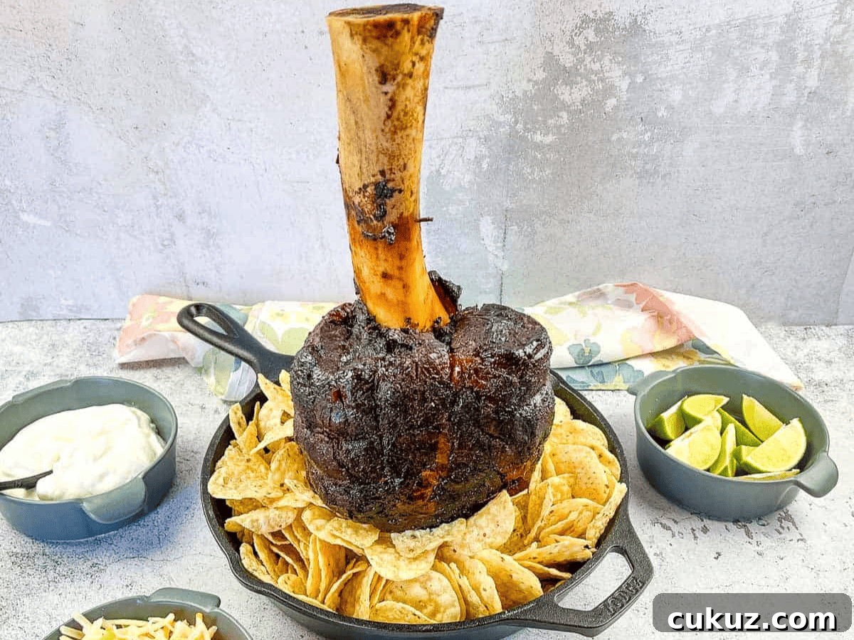 A large roasted meat shank on a bone, displayed upright in a skillet surrounded by nachos, with bowls of lime wedges and sauce nearby.