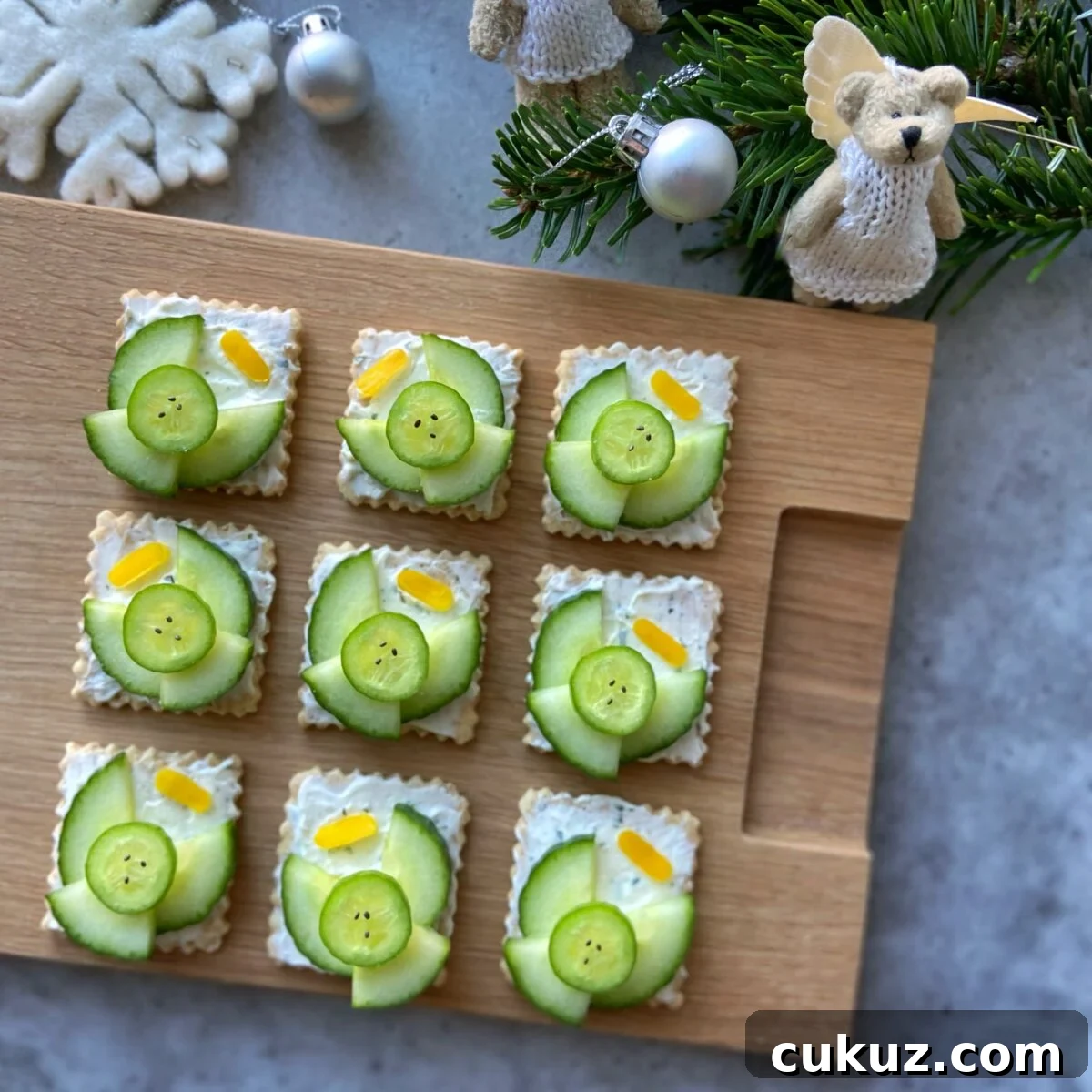 Nine square crackers topped with cream cheese, cucumber slices, and small yellow pepper halos. They are arranged on a wooden board with holiday decorations nearby.