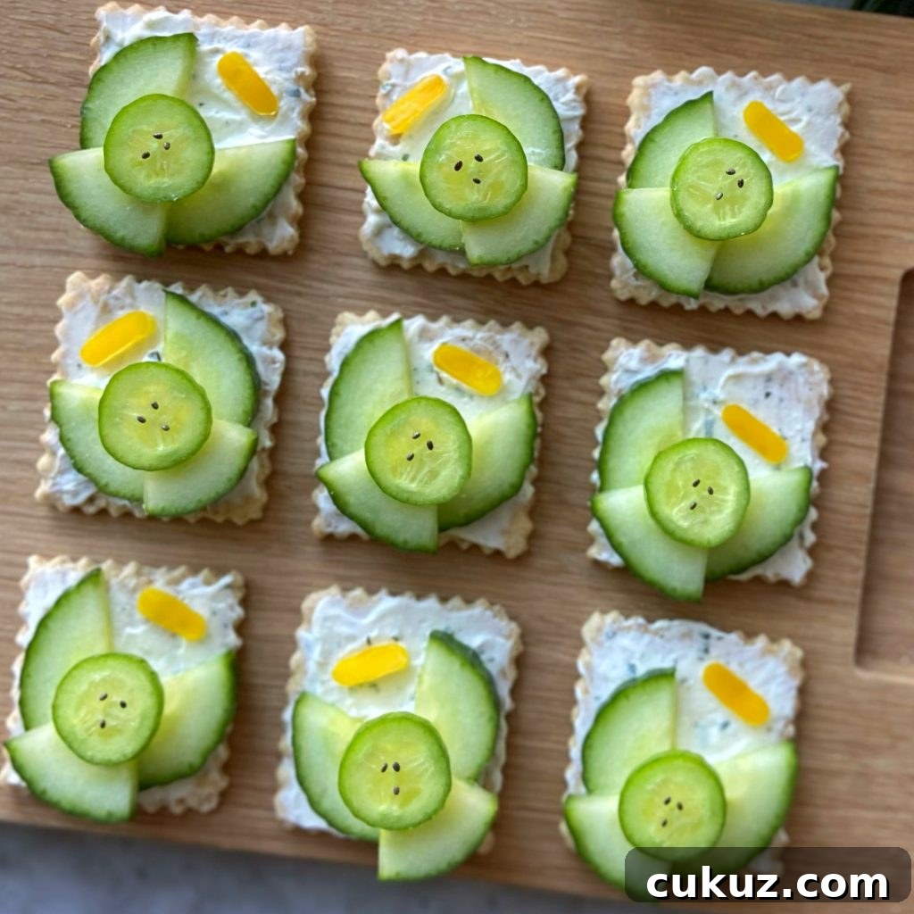 Closeup of nine crackers topped with a creamy spread, cucumber slices arranged as angels with small yellow pepper halos. They are displayed neatly on a wooden board.