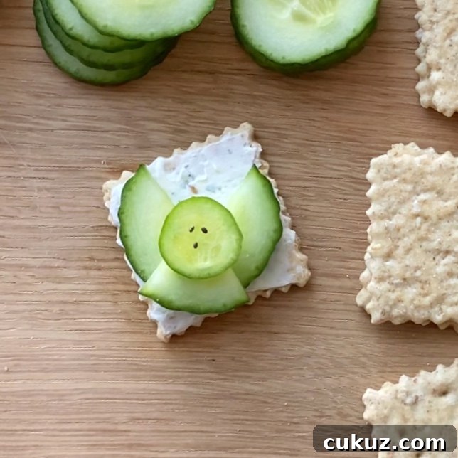 A square cracker with a layer of cream cheese, topped with cucumber slices arranged to resemble an angel.