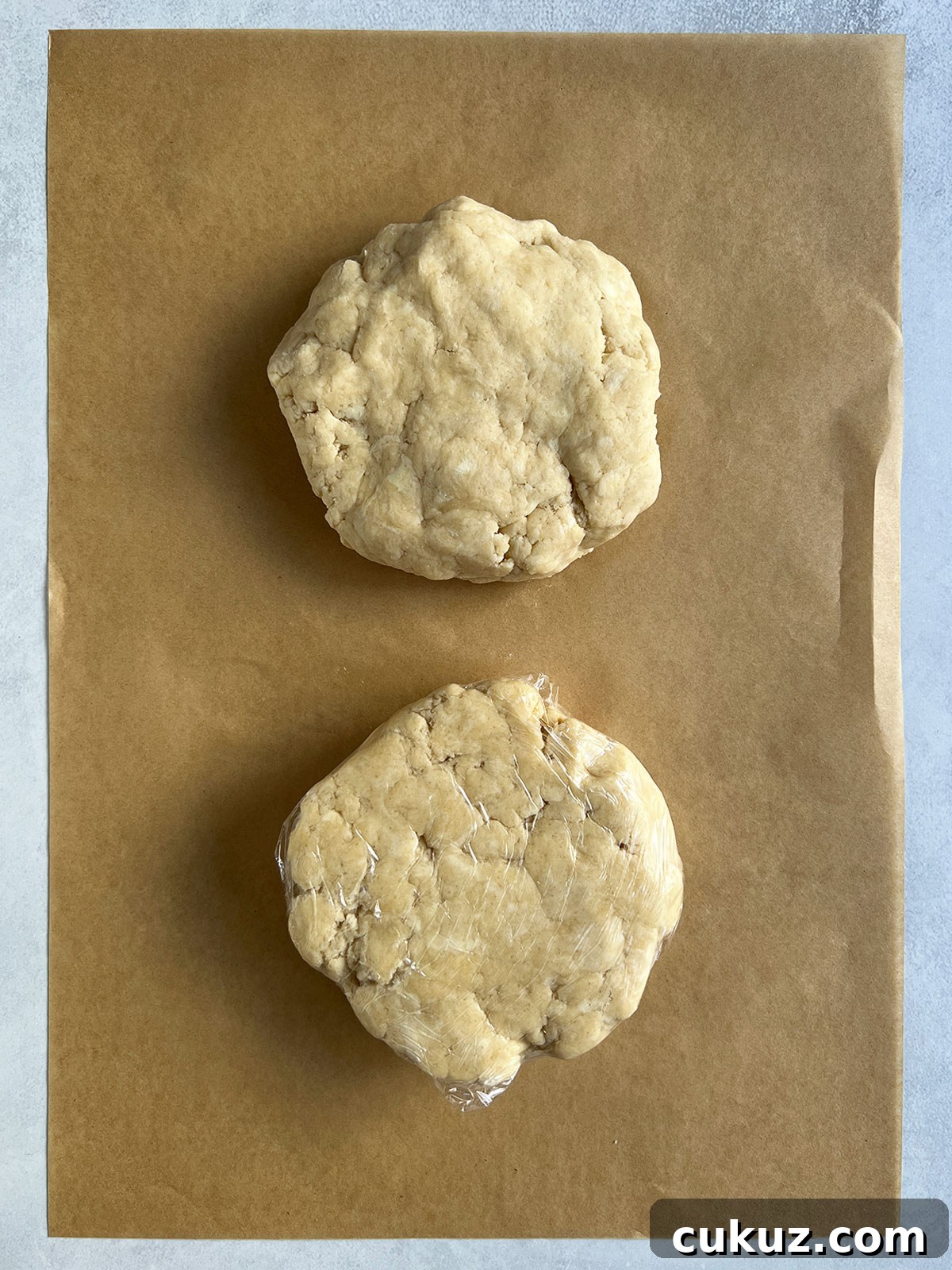 Two disks of pie dough on a piece of parchment - one wrapped in plastic wrap and one not.