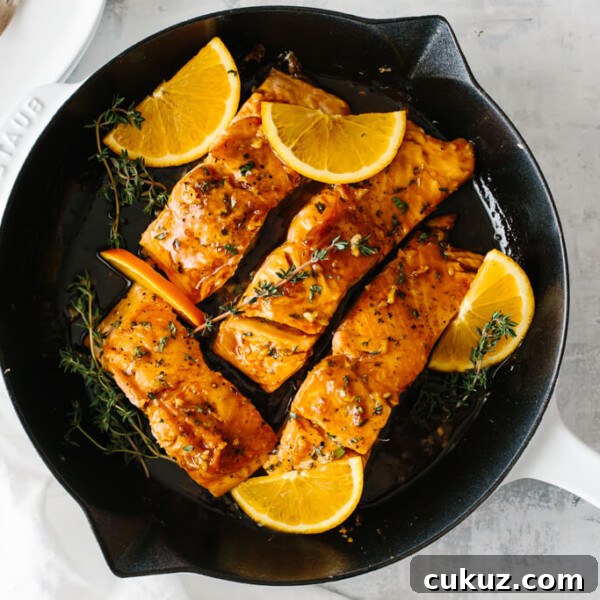 Four salmon filets in a pan with orange slices and thyme on top.