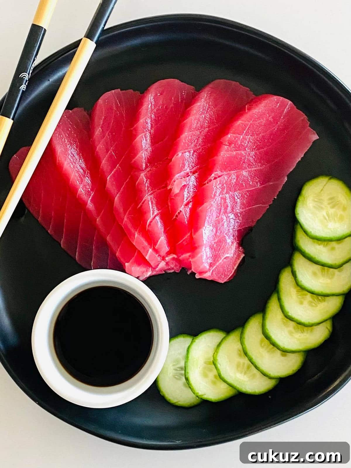 tuna sashimi on plate with soy sauce, sliced cucumbers and chopsticks.
