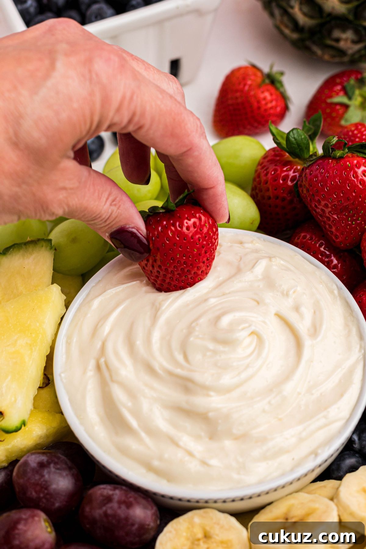 A hand dipping a bright red strawberry in a bowl of white creamy dip, surrounded by fruits.